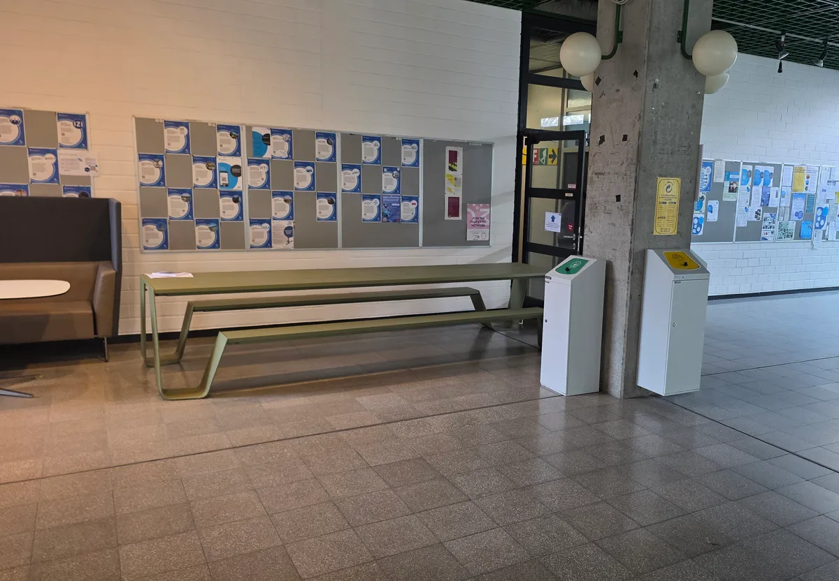 Grüner Tisch mit Bänken vor der weißen Wand mit Aushängen im Gebäude Pfaffenwaldring 9, vor V9.02.